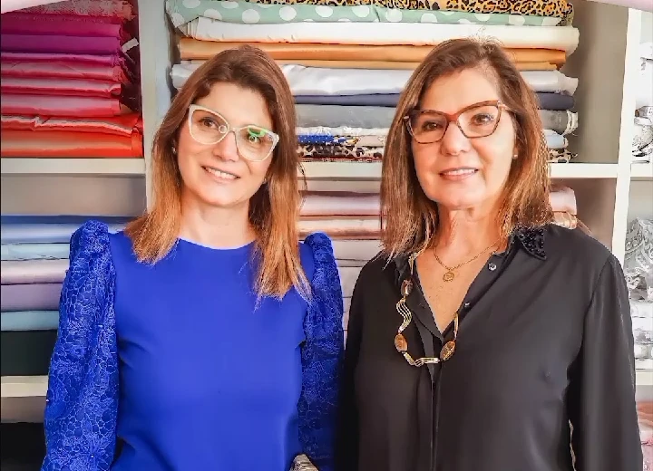 Anita e Leticia, mãe e filha, no ateliê, em frente às prateleiras de tecidos
